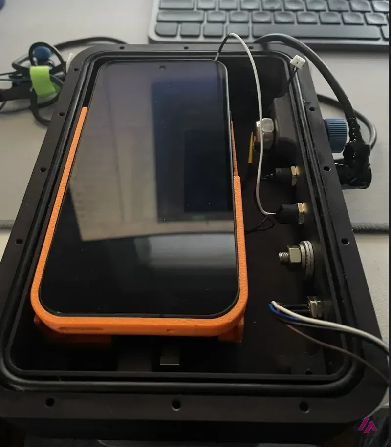 A Google Pixel 9 with an orange frame mounted inside an open black waterproof housing, with cables and connectors on one side